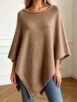 A woman wearing a loose, scoop-neck knit poncho sweater in a warm taupe-brown color, paired with blue jeans, standing in a brightly lit room with white walls and wood floors.