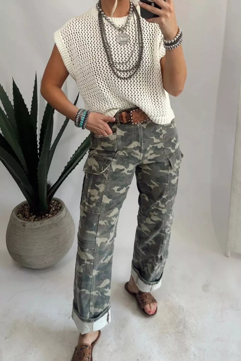 Person wearing a white sleeveless top and camouflage pants, standing indoors with a plant in the background.