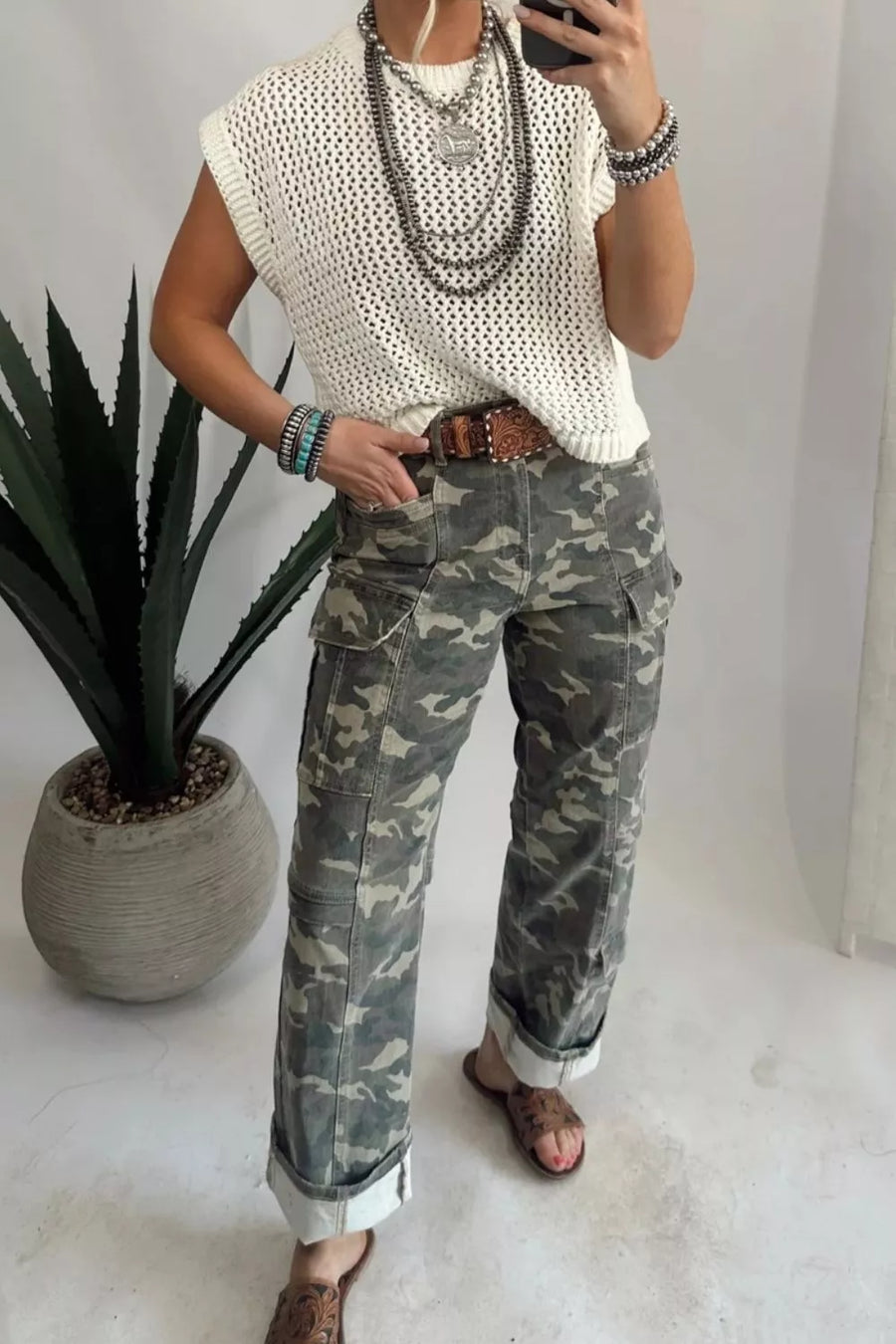 Person wearing a white sleeveless top and camouflage pants, standing indoors with a plant in the background.