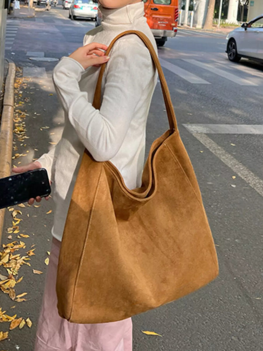 A woman on a city street carries a large tan suede-like shoulder bag. The bag has a slouchy, open-top design with a single wide shoulder strap and is shown in a real outdoor environment with cars and road markings visible.