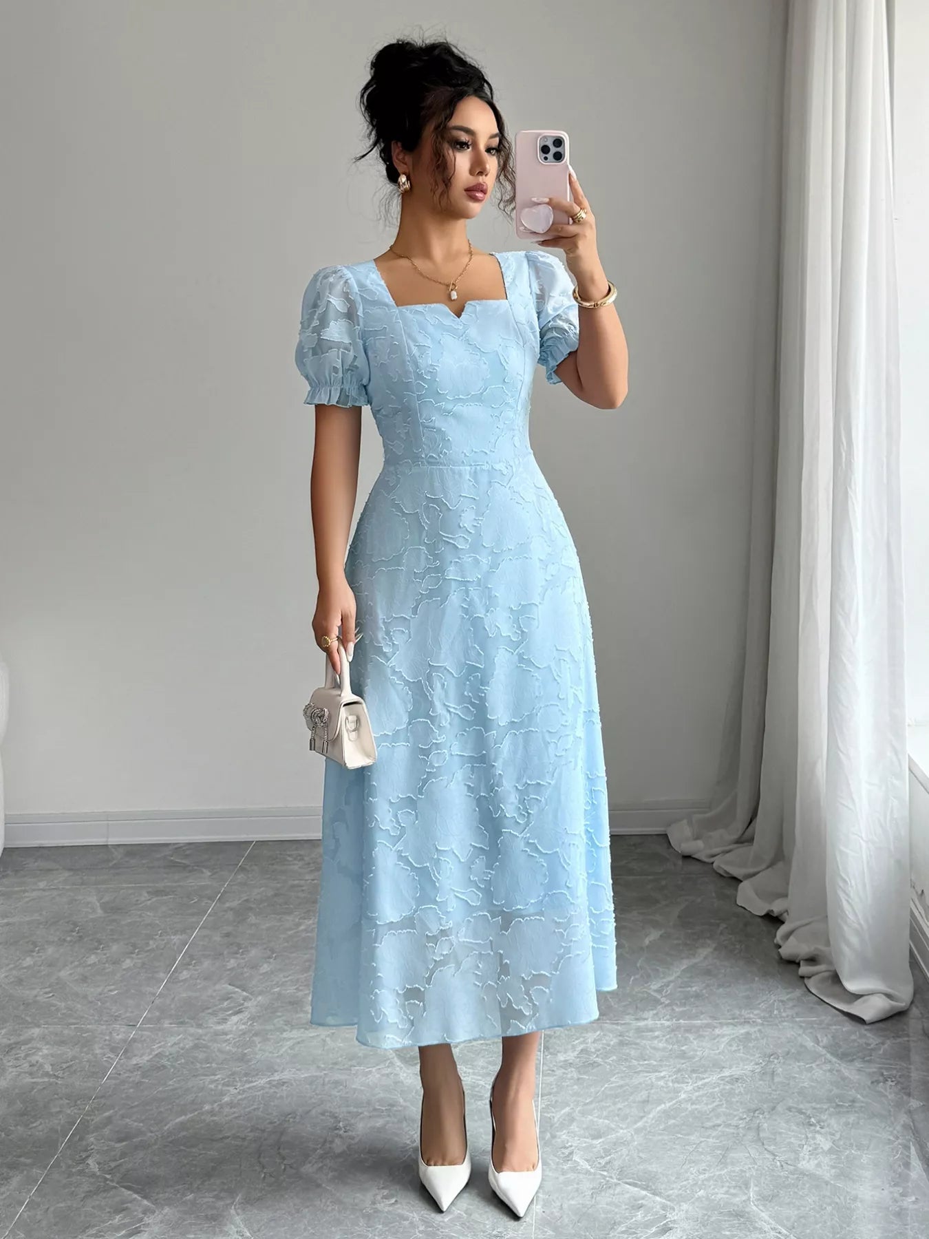 Woman in a light blue lace dress taking a selfie in a room with white curtains.