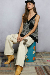 Woman sitting on a colorful stool wearing a black and white patterned top and beige pants.