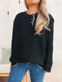 Person wearing a dark green sweater with button details and blue jeans, standing against a light-colored wall.