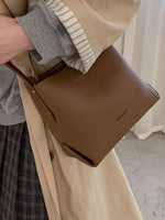 Brown leather handbag held by a person wearing a beige coat and plaid skirt.