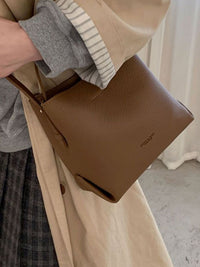 Brown leather handbag held by a person wearing a beige coat and plaid skirt.