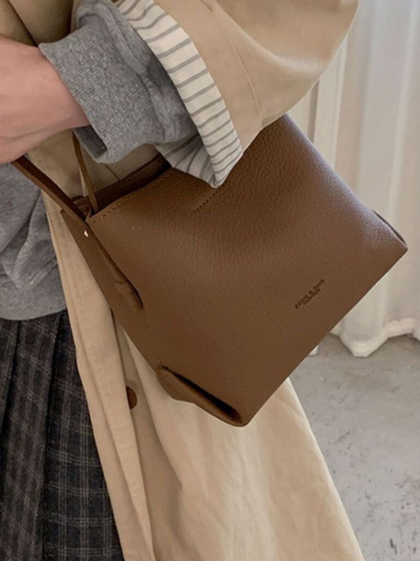 Brown leather handbag held by a person wearing a beige coat and plaid skirt.