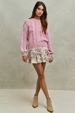 Woman wearing a pink cable knit sweater and floral skirt against a neutral background