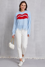 Woman wearing a light blue sweater with a red design, white pants, and holding a white handbag on a gray background