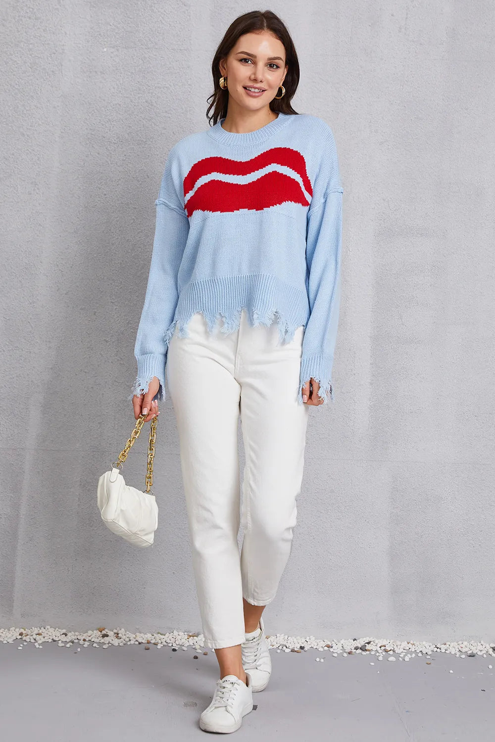 Woman wearing a light blue sweater with a red design, white pants, and holding a white handbag on a gray background