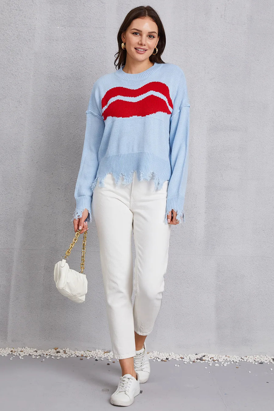 Woman wearing a light blue sweater with a red design, white pants, and holding a white handbag on a gray background