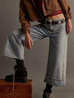 Person wearing a brown jacket, red top, and blue jeans sitting on a suitcase against a gray background