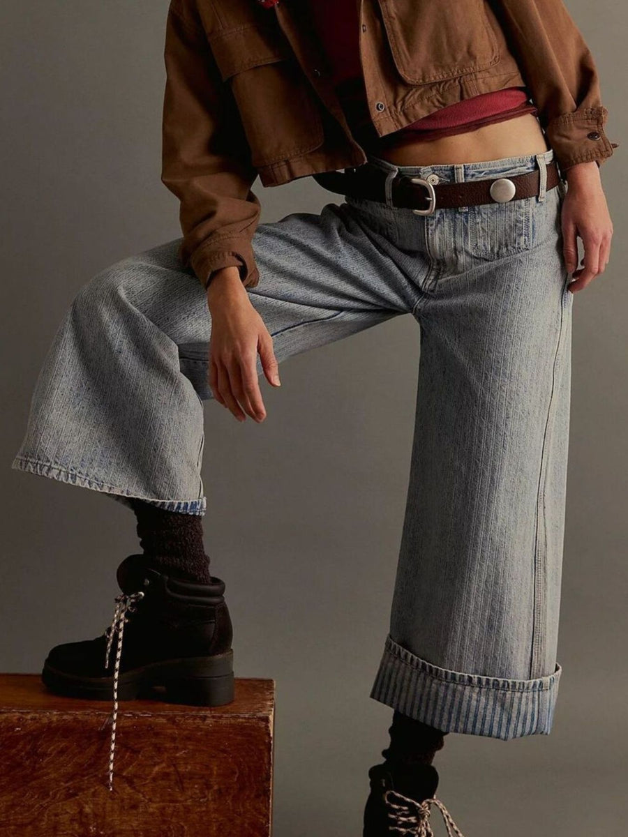 Person wearing a brown jacket, red top, and blue jeans sitting on a suitcase against a gray background