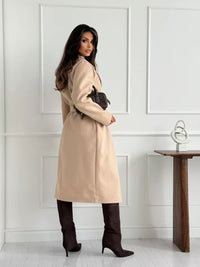 Woman wearing a beige coat in a minimalistic room with a white wall and wooden table.