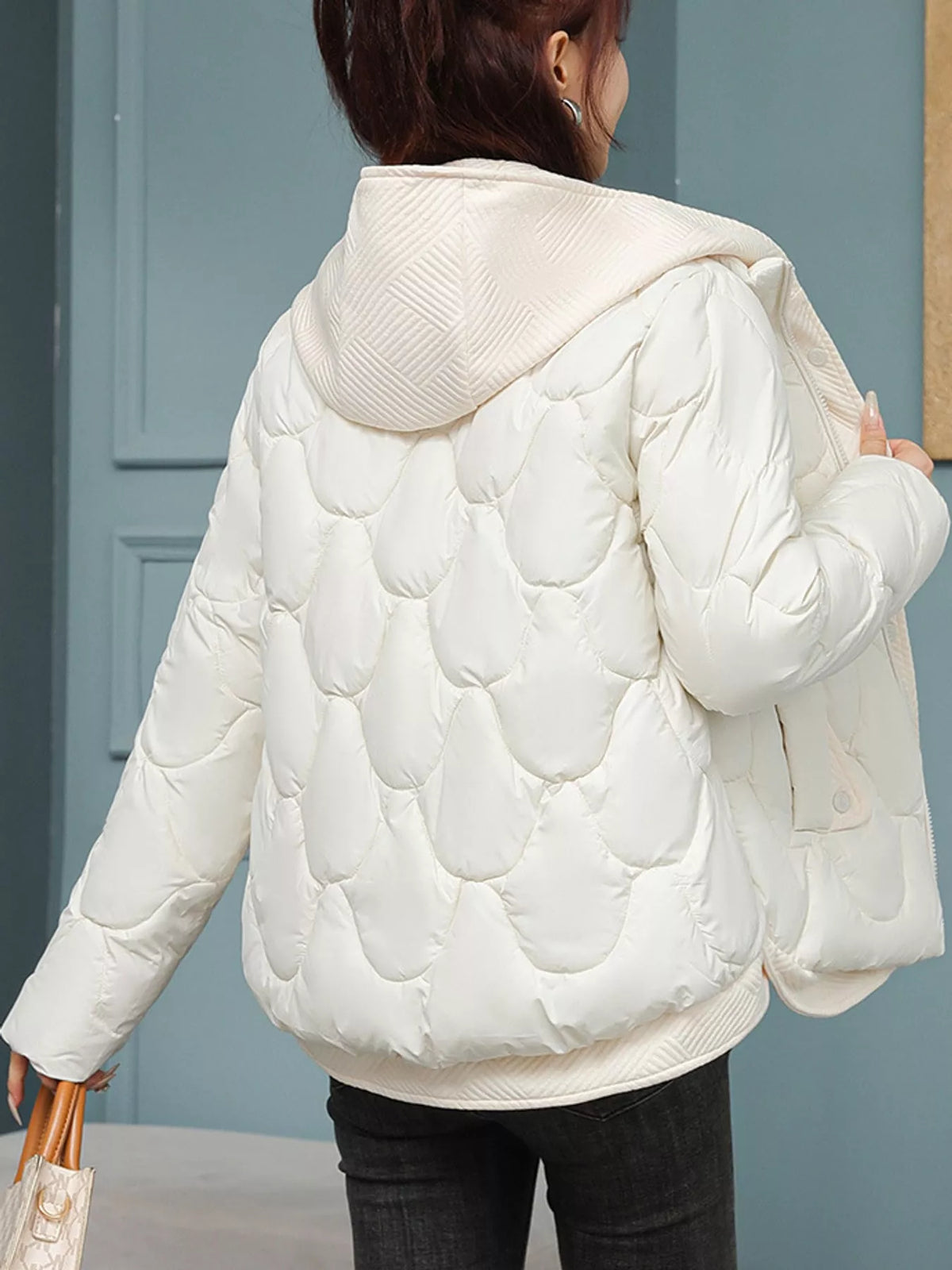 A woman shown from the back wearing a cream-colored quilted puffer jacket with a hood, paired with black pants, standing in front of a blue paneled wall.