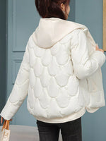 A woman shown from the back wearing a cream-colored quilted puffer jacket with a hood, paired with black pants, standing in front of a blue paneled wall.