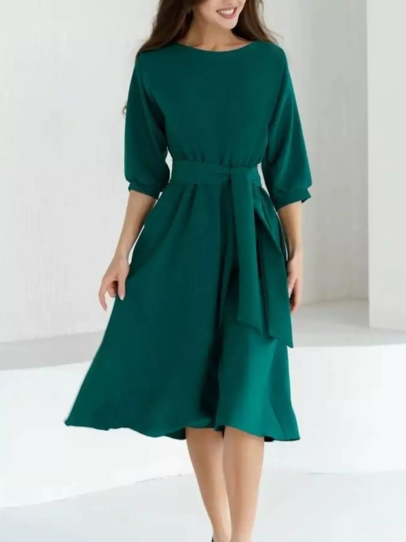 A woman wearing a dark green midi dress with three-quarter sleeves, a round neckline, and a matching fabric tie belt at the waist, shown against a bright white studio background.