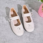 A pair of off-white Mary Jane style shoes with decorative silver-tone embellishments on the toes and buckle straps, displayed on a gray textured surface.