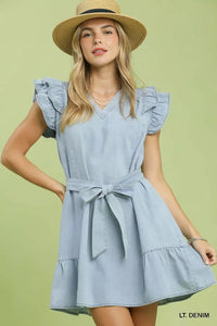 Woman wearing a light blue dress with ruffled sleeves and a belt, standing against a light green background.