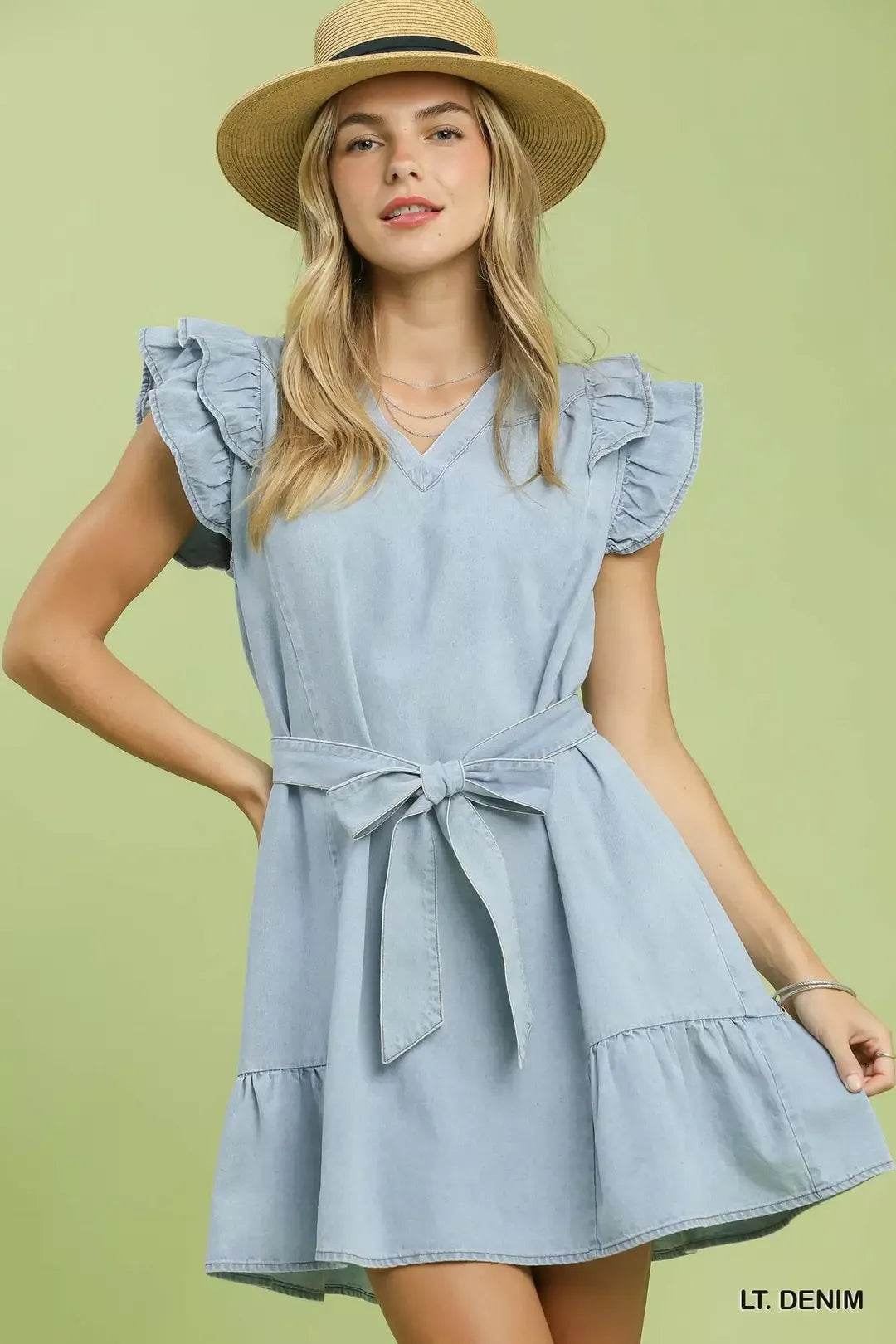 Woman wearing a light blue dress with ruffled sleeves and a belt, standing against a light green background.