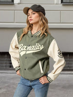 A woman wearing an olive green and cream varsity jacket with 'Baronity' written across the chest, paired with blue jeans and a dark cap, standing against a gray building wall.