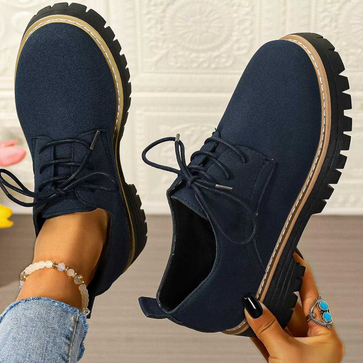 Navy blue shoes with thick soles held by a person against a textured white wall.
