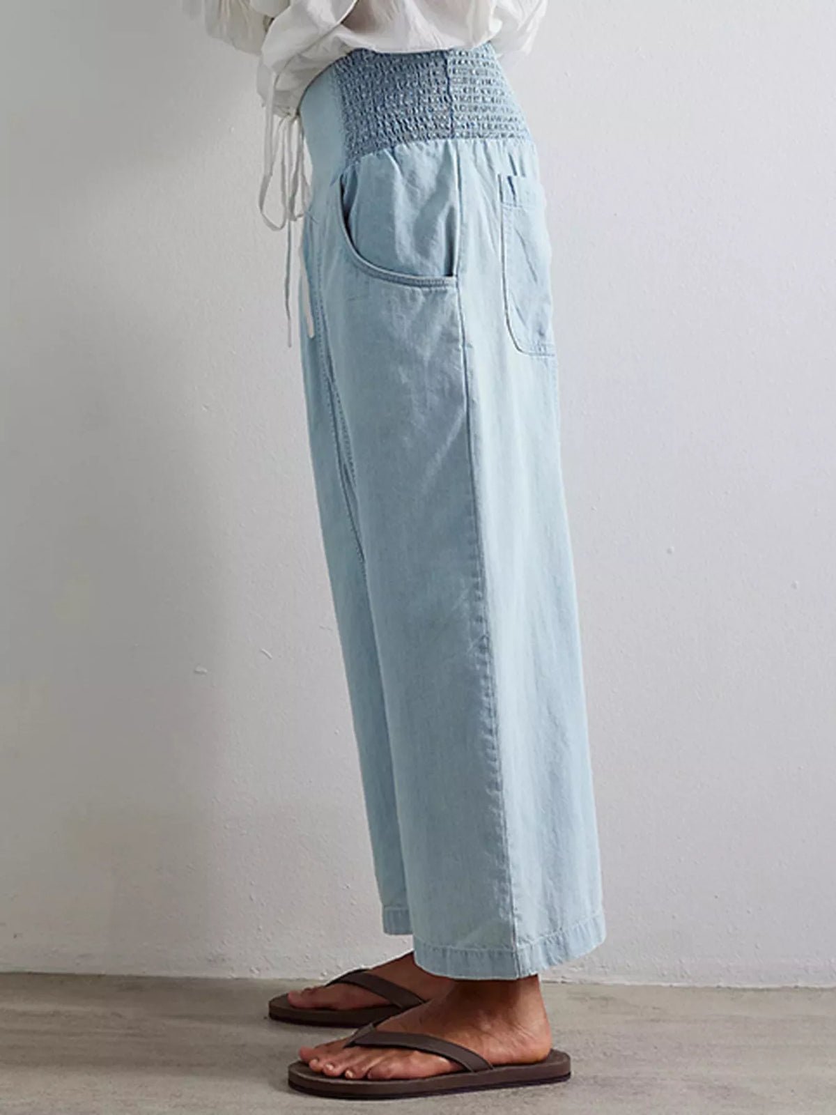 A person wearing light blue wide-leg cropped pants with a smocked waistband and side pockets, paired with a white top and dark brown flip-flop sandals, standing against a plain light gray wall.