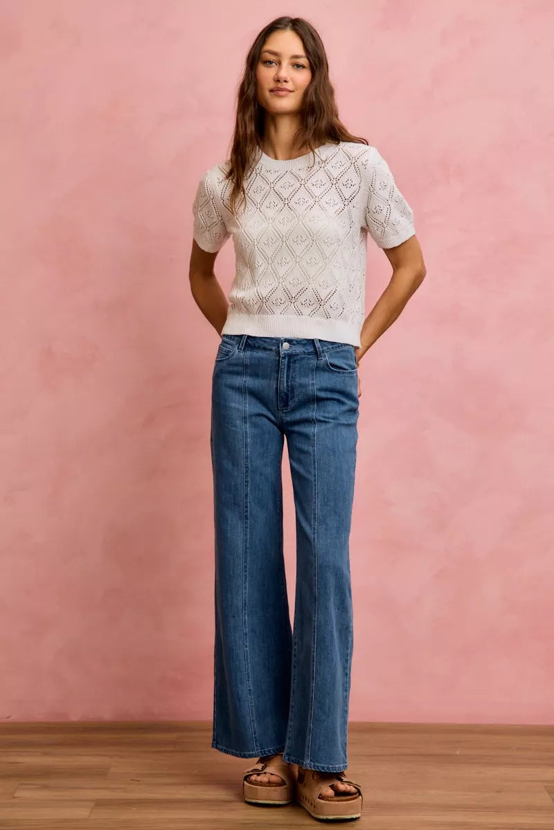 Woman wearing a white patterned top and blue jeans against a pink wall.