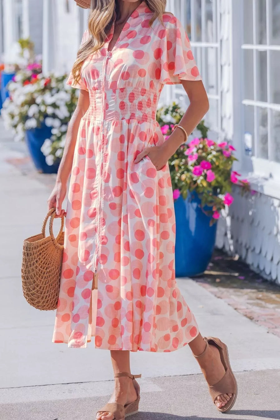 Woman wearing a pink polka dot dress with a straw bag in an outdoor setting.