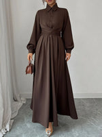 Woman wearing a long brown dress holding a matching handbag against a neutral background