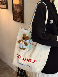 Person holding a white tote bag with a teddy bear and text, wearing a black hoodie.
