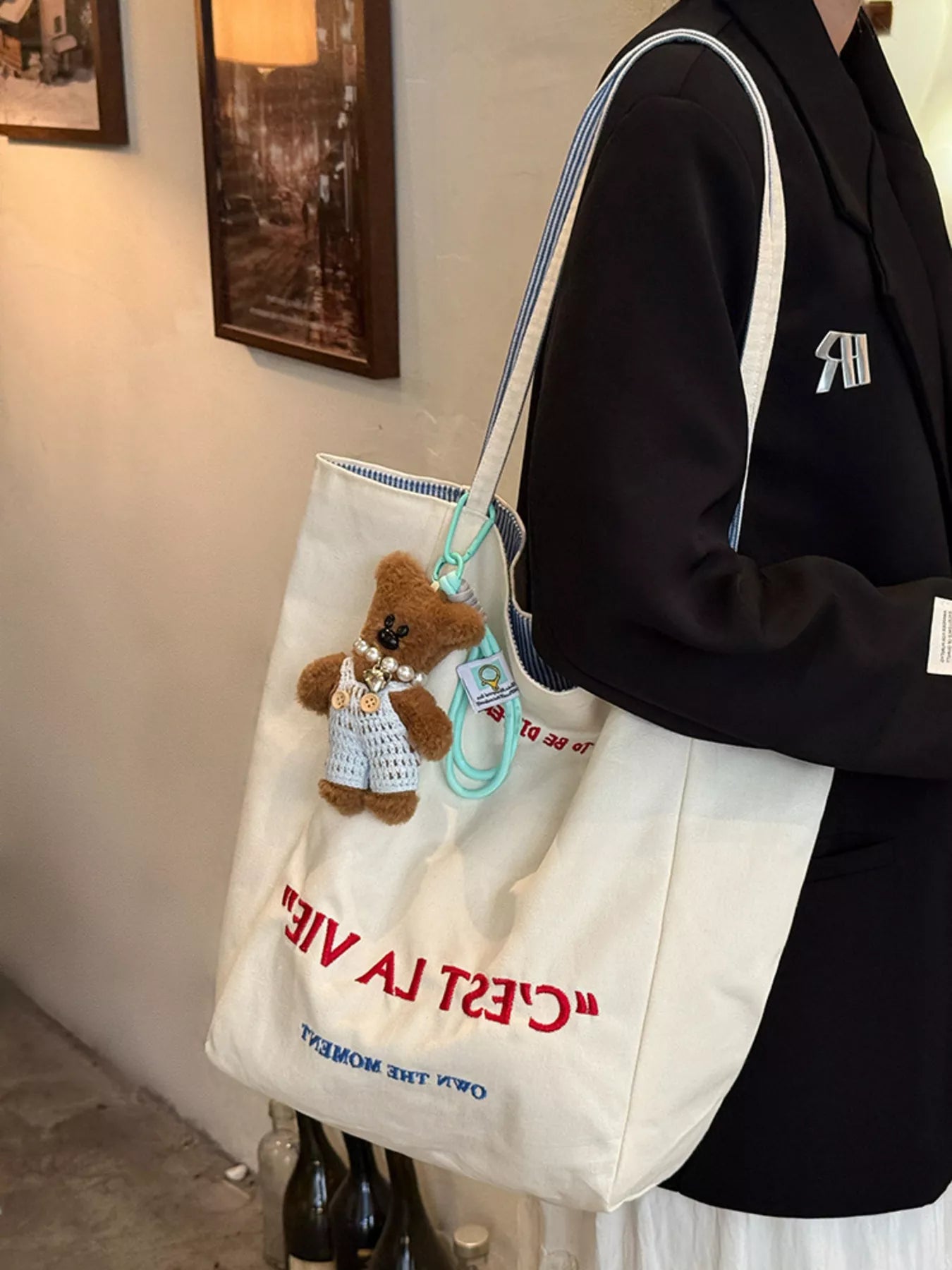 Person holding a white tote bag with a teddy bear and text, wearing a black hoodie.