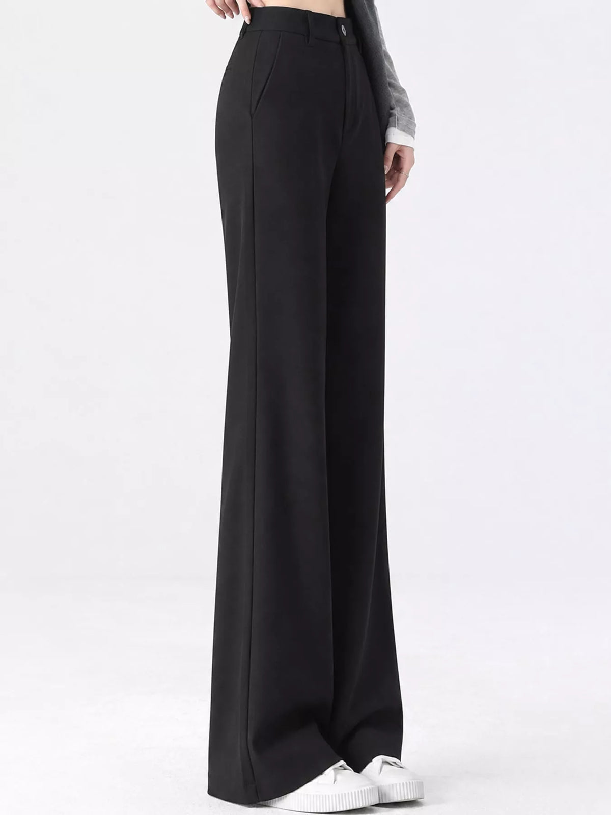 A person wearing black wide-leg trousers with a high waist, paired with white sneakers and a gray sleeve, shown from the waist down against a plain white studio background.