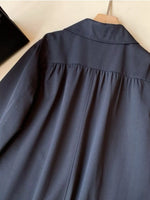 A dark slate blue long-sleeve shirt with a gathered waist seam, hanging on a wooden hanger against a light beige wall.