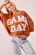 Person wearing a brown 'GAMEDAY' sweatshirt with light blue jeans on a plain background