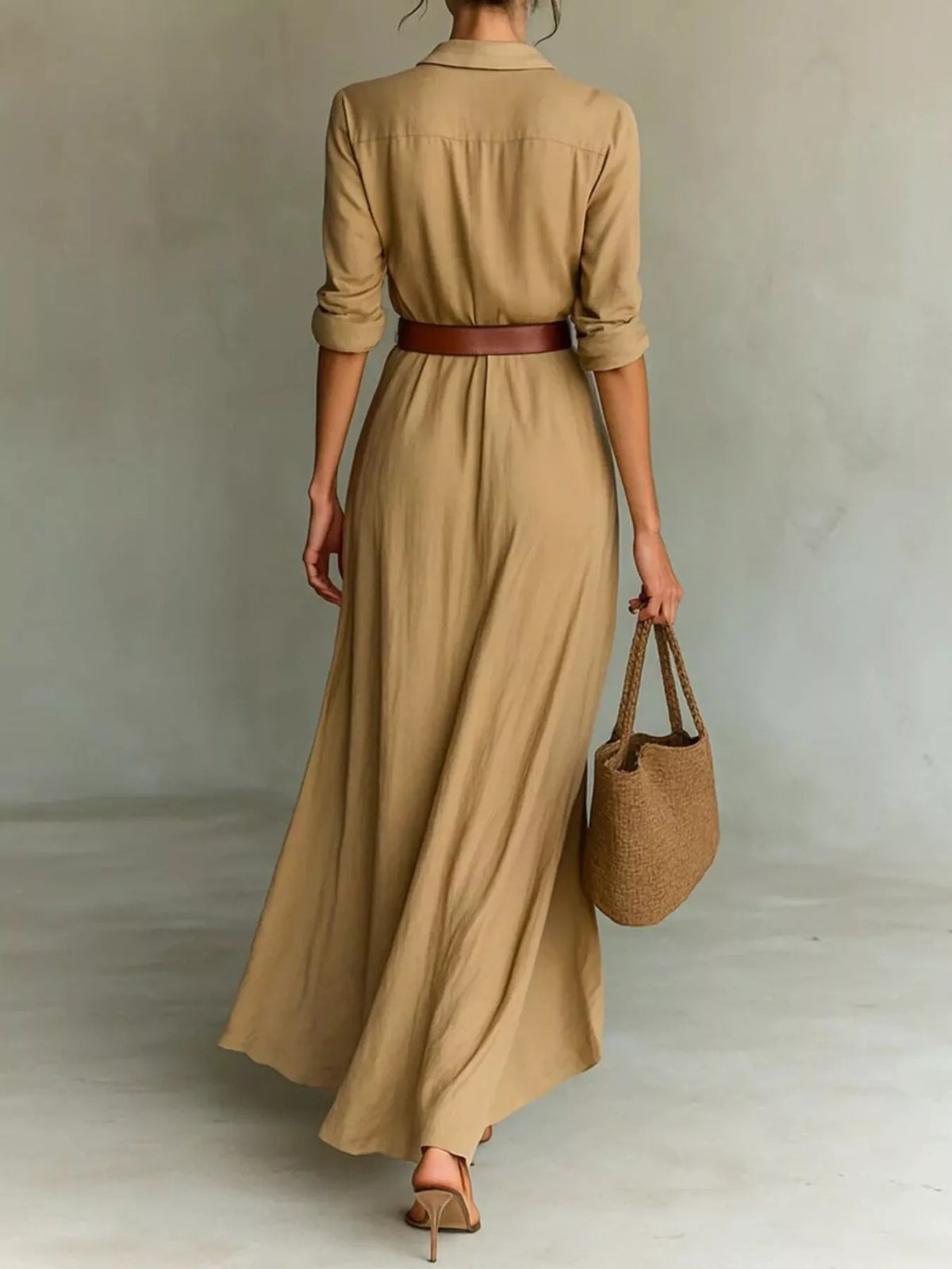 A woman shown from the back wearing a tan maxi shirt dress with three-quarter sleeves and a thin brown belt, holding a woven straw bucket bag against a neutral studio backdrop.