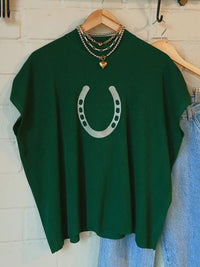 Green sweater with a horseshoe design on a hanger against a white wall.