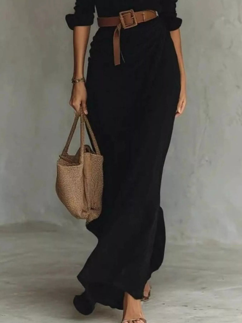 Person wearing a black outfit with a brown belt, holding a woven bag against a neutral background