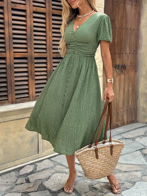 Woman wearing a green dress holding a straw bag in front of a wooden door.
