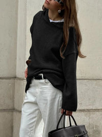 Person wearing a black oversized sweater and white pants, holding a black bag against a light-colored wall.