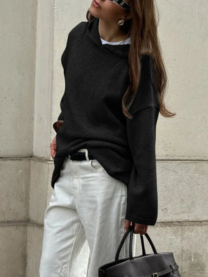 Person wearing a black oversized sweater and white pants, holding a black bag against a light-colored wall.