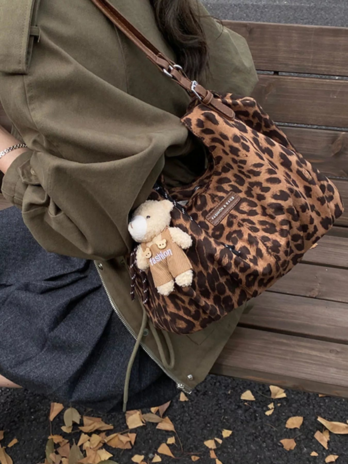 Person holding a leopard print bag with a teddy bear attached, sitting on a wooden bench.