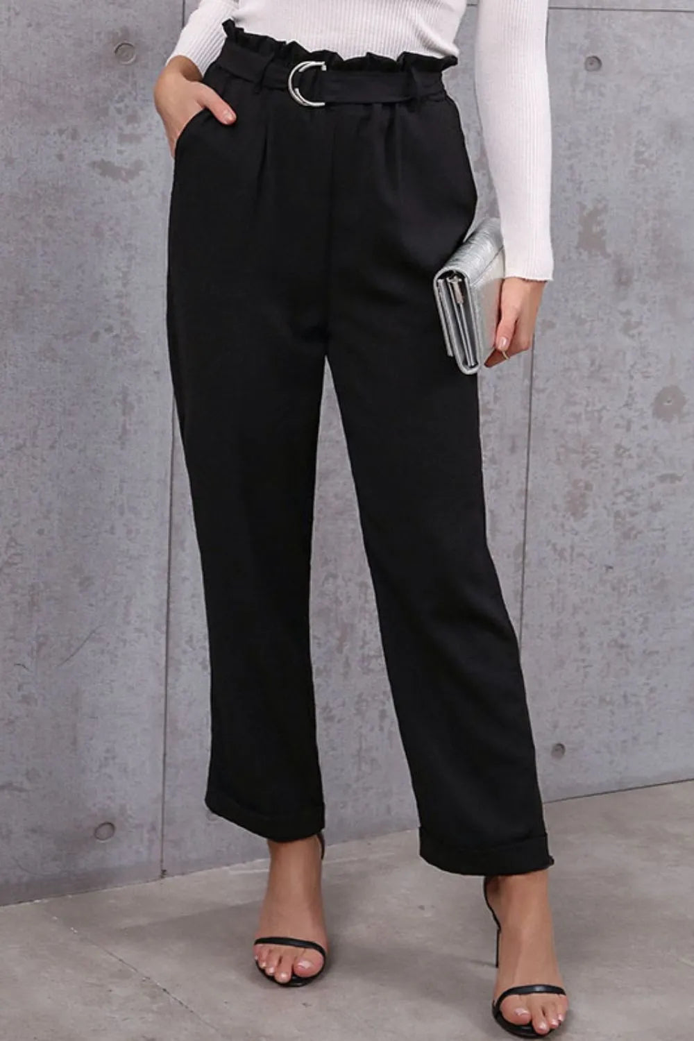 Person wearing black high-waisted pants with a belt against a gray concrete wall.