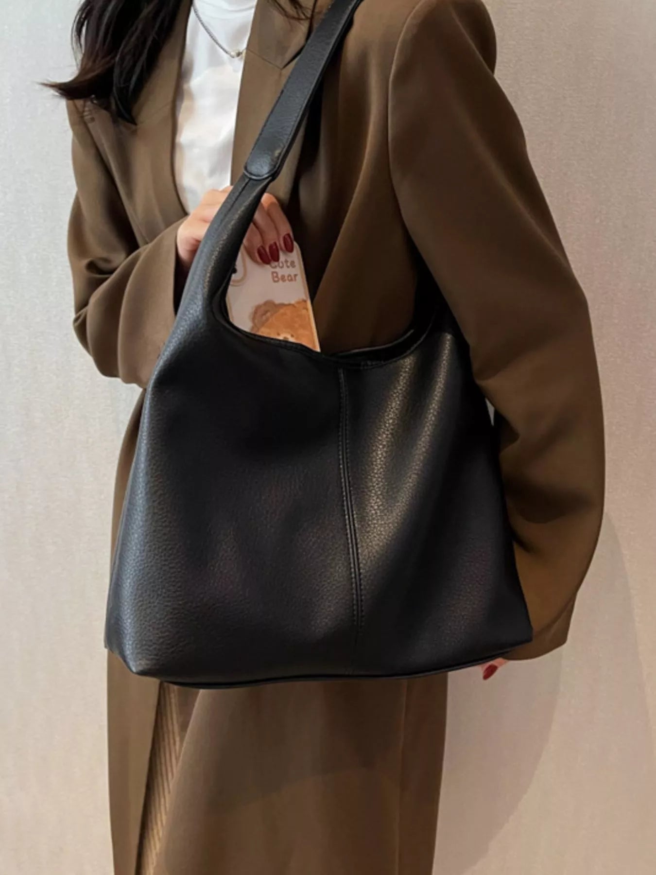 Person wearing a brown coat holding a black leather handbag against a neutral background
