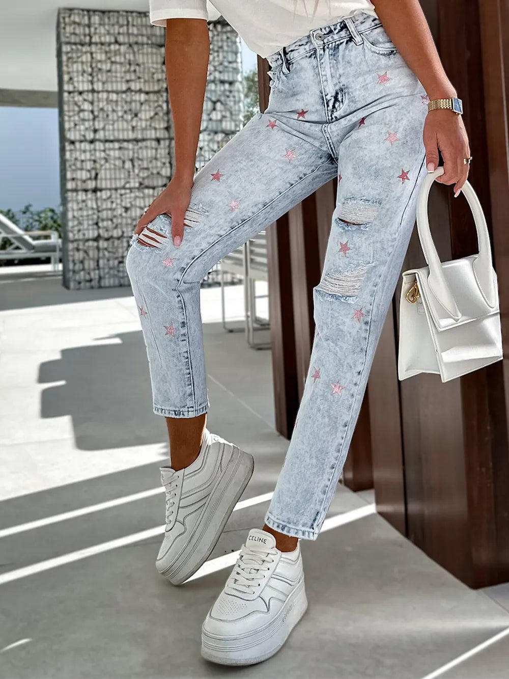 Person wearing light blue jeans with floral embroidery and white sneakers, holding a white handbag.