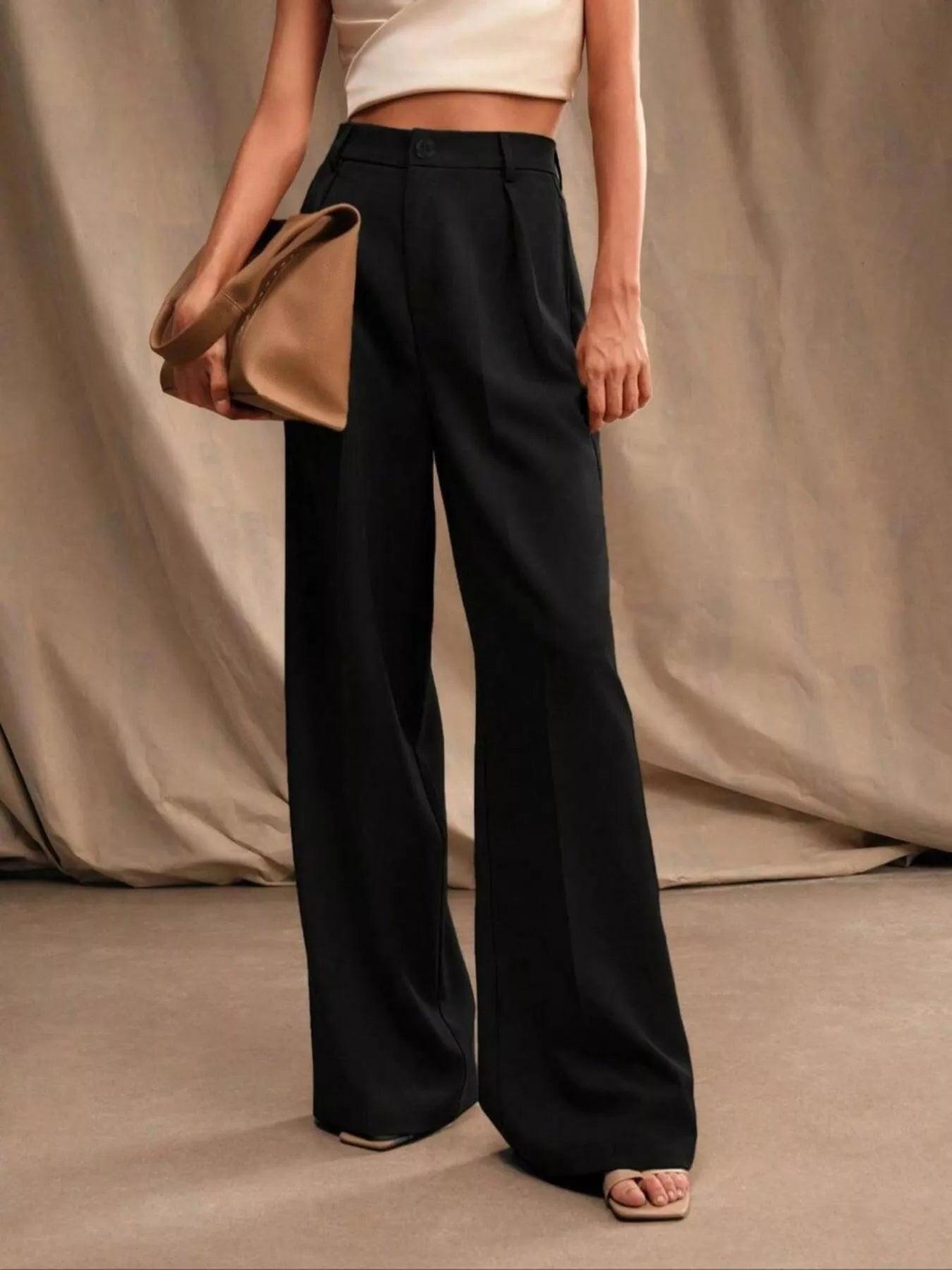 Person wearing black wide-leg pants holding a beige clutch against a neutral background
