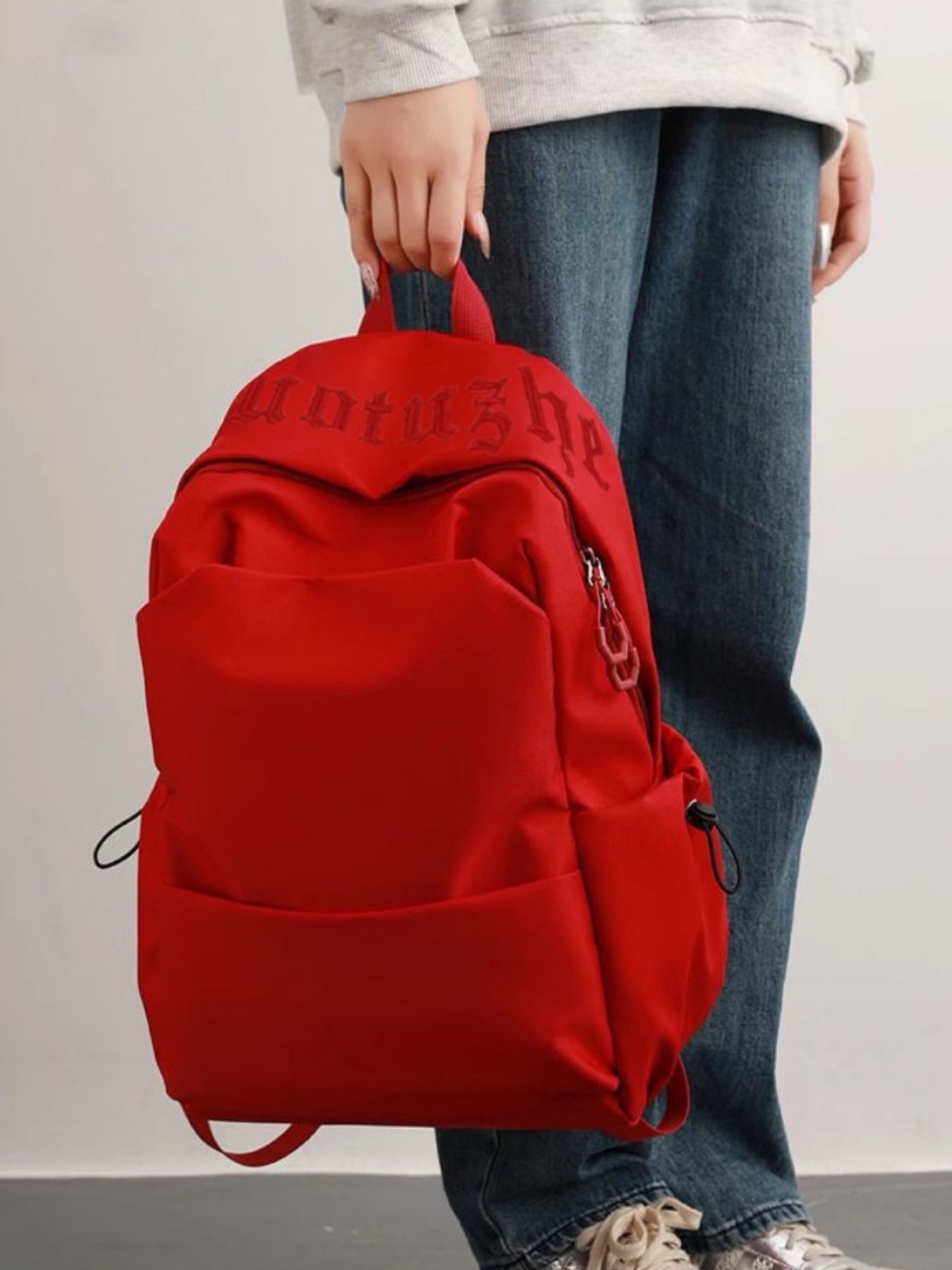 Red backpack held by a person wearing jeans and a light-colored top on a plain background