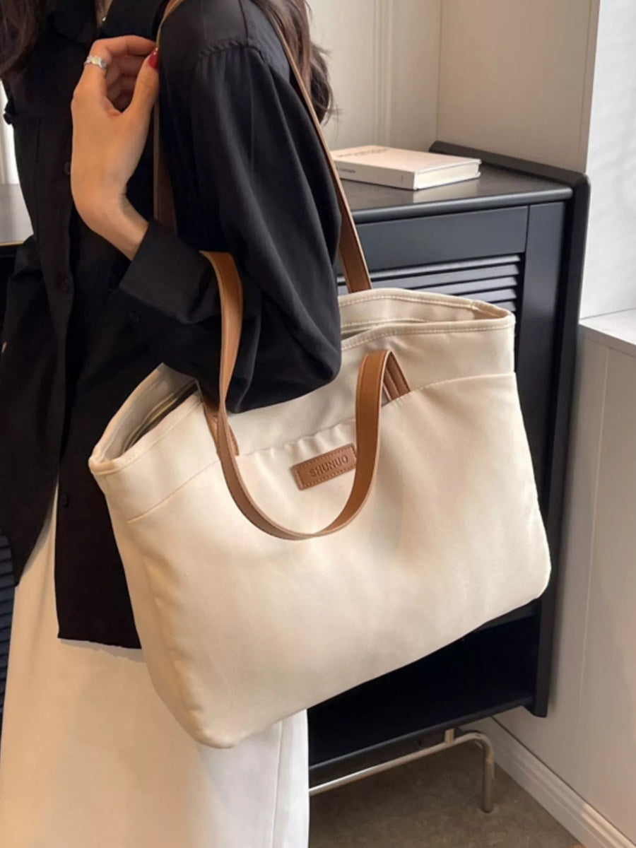 Beige tote bag with brown handles held by a person in a black outfit.
