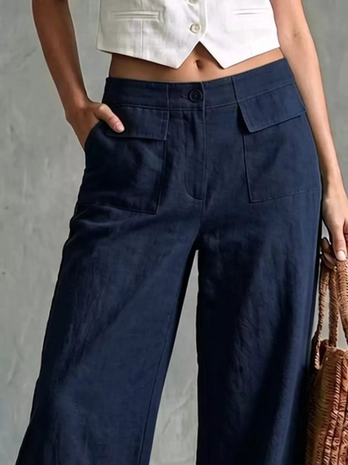 A person wearing dark navy wide-leg trousers with a white cropped top, standing against a textured gray wall and holding a woven brown bag.