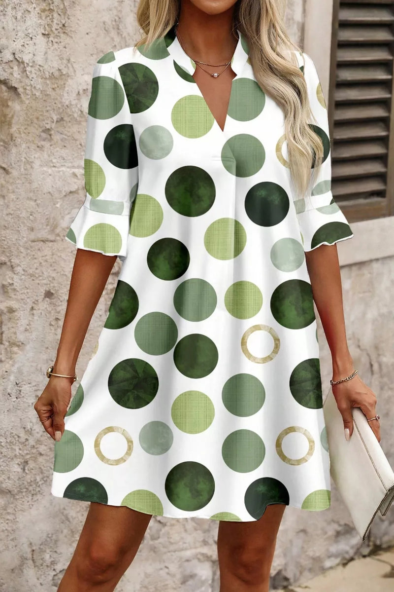 Woman wearing a white dress with green polka dot pattern against a stone wall.