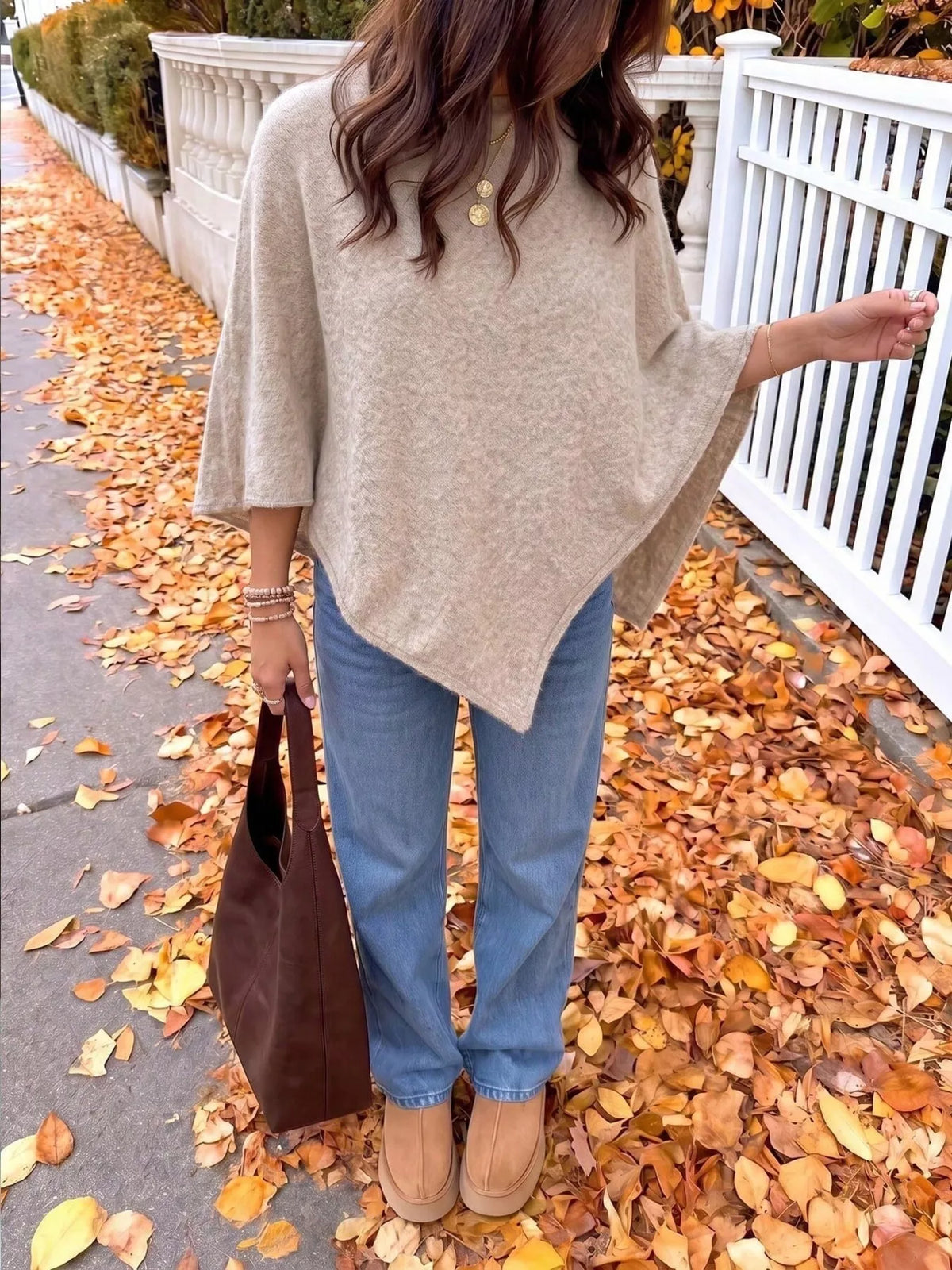 A woman wearing a beige knit poncho sweater, blue jeans, and tan boots stands on a sidewalk covered with fallen autumn leaves. She is holding a dark brown leather tote bag, with a white fence in the background.